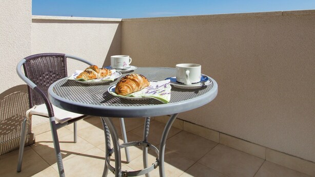 Una mesa redonda de metal con una taza blanca y dos croissants en un balcón bajo un cielo azul despejado.