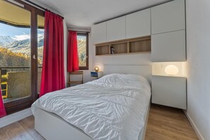 A bedroom with a bed, a nightstand, a lamp, a window with curtains, and a view of mountains. - 100m from ski slopes, 1st floor, view mountain, balcony, tv, 38m², Les Orres (Les Orres)