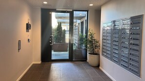 Un couloir avec un sol carrelé, une plante en pot, une porte avec un panneau de verre et une boîte aux lettres fixée au mur.