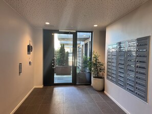 Un couloir avec un sol carrelé, une plante en pot, une porte avec un panneau de verre et une boîte aux lettres fixée au mur.