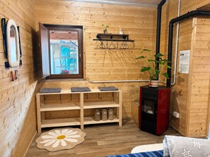 A wooden cabin interior with a window, a shelf with jars, a fireplace, and a bed.