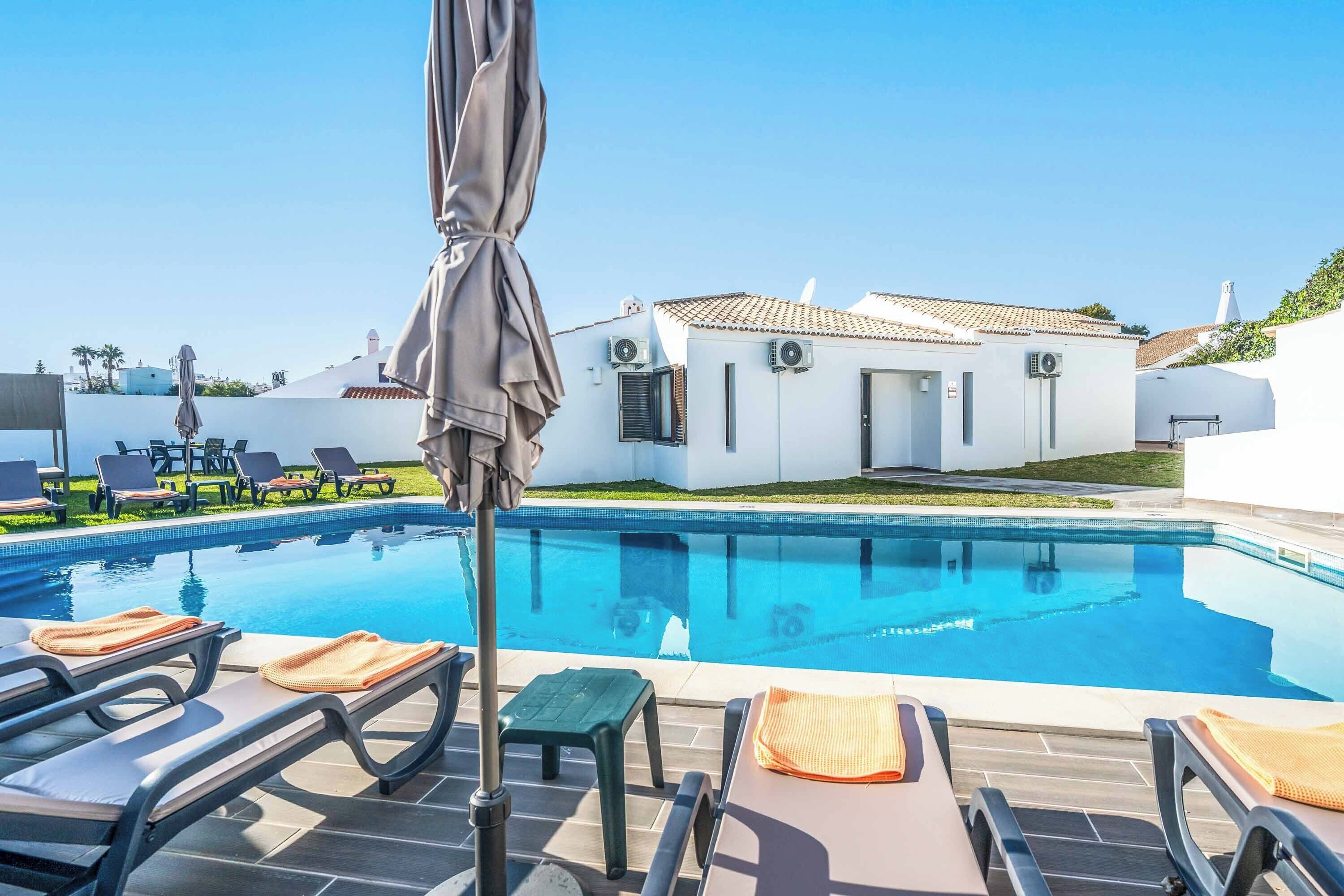 A swimming pool with lounge chairs and umbrellas, a white building with a tiled roof, and a clear blue sky.
