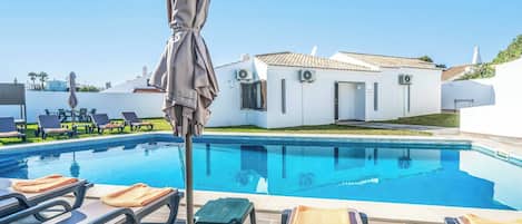 A swimming pool with lounge chairs and umbrellas, a white building with a tiled roof, and a clear blue sky.