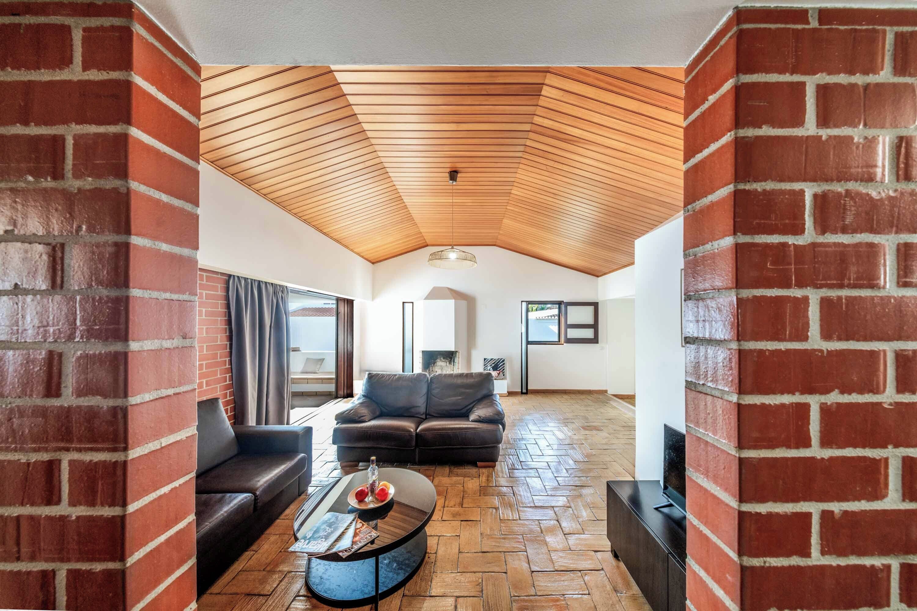 A spacious living area with a herringbone floor pattern, a wooden slatted ceiling, and a brick wall.