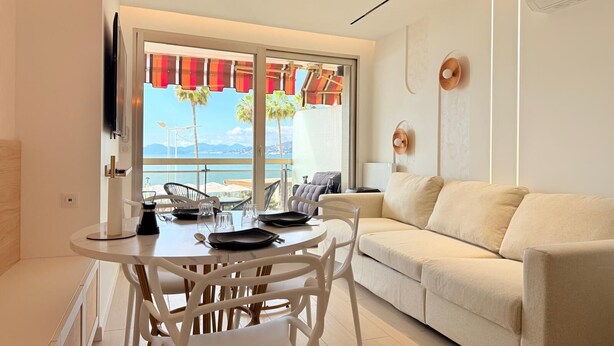 En moderne stue med balkon, en hvid sofa, et spisebord med stole og et fjernsyn ophængt på væggen.