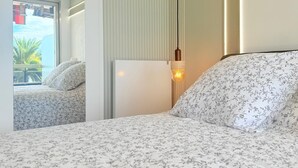 Et hotelværelse med en seng, et natbord, en lampe og et vindue med udsigt til palmer.