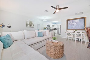 Ein modernes Wohnzimmer mit einem Ecksofa, einem runden gewebten Couchtisch, einem Essbereich mit weißen Stühlen und einem Deckenventilator.
