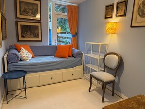 Una habitación con cama, una silla, una mesita, una ventana con cortinas y cuadros en la pared.