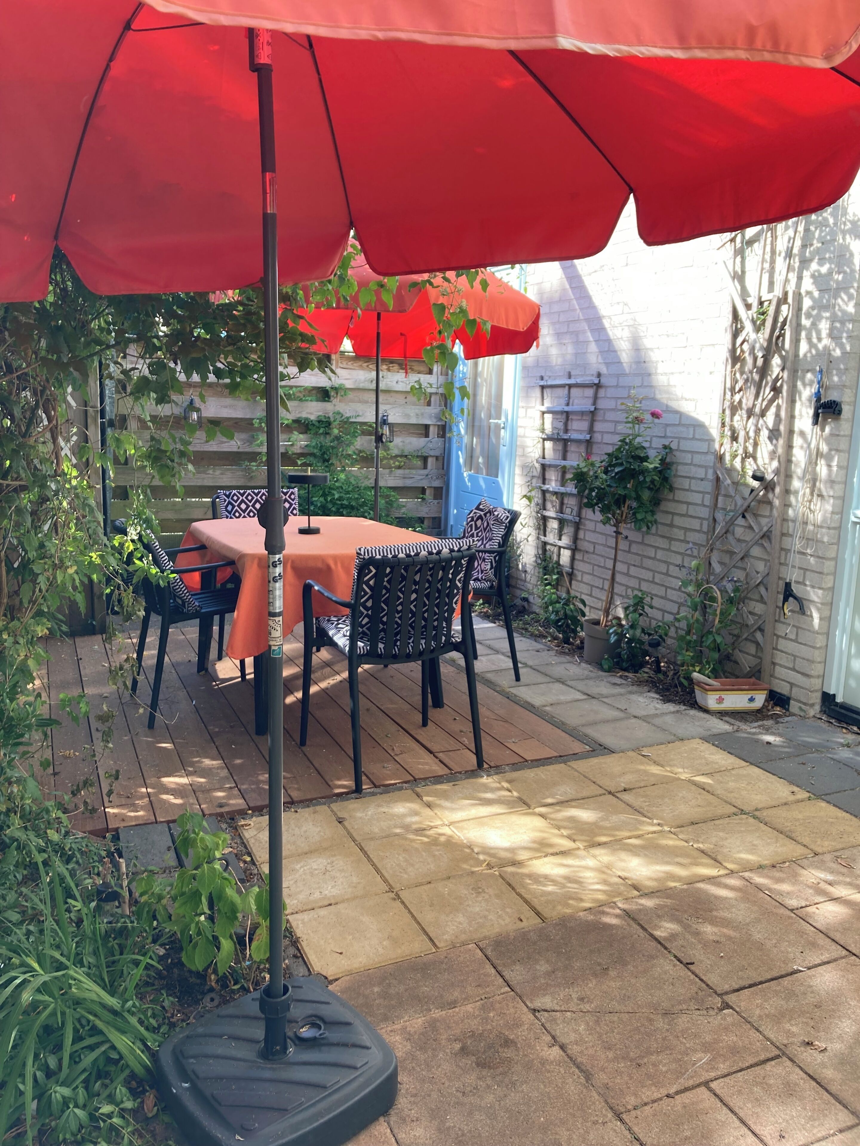 Een terras buiten, met een tafel, stoelen, een rode parasol en een bakstenen muur.