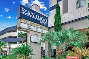 Ein mehrstöckiges Gebäude mit einem auffälligen Schild für das Beach Colony Resort, inklusive Annehmlichkeiten wie einem Café, einer Lounge und einem Verkaufsbüro für Apartments mit Meerblick.