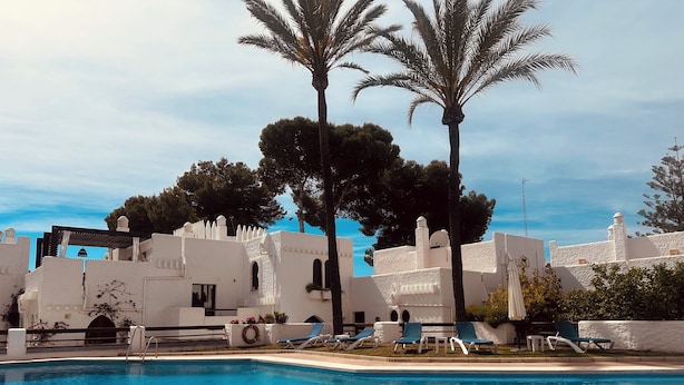 A pool with lounge chairs, palm trees, and a white building with arches. - Penthouse in Puerto Banús (Marbella)