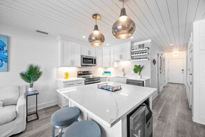 A modern kitchen with white cabinets, a central island, pendant lighting, and a dining area with a white table and blue chairs. - Seagull Villas 4 | Total Remodel | Private Beach | Nearby Dining | Beach Gear (Panama City Beach)