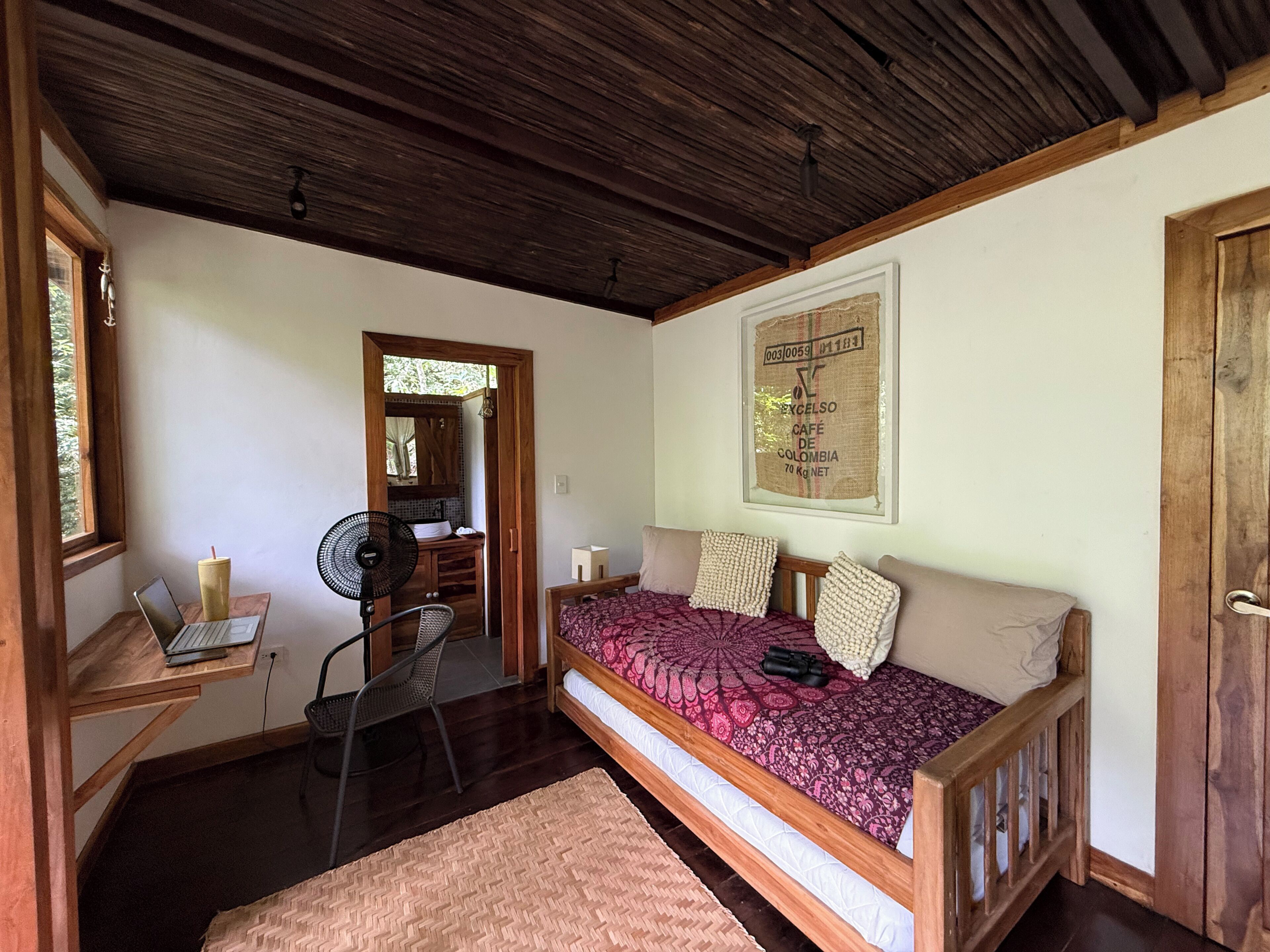 A room with a bed, a desk with a laptop, a fan, and a door.