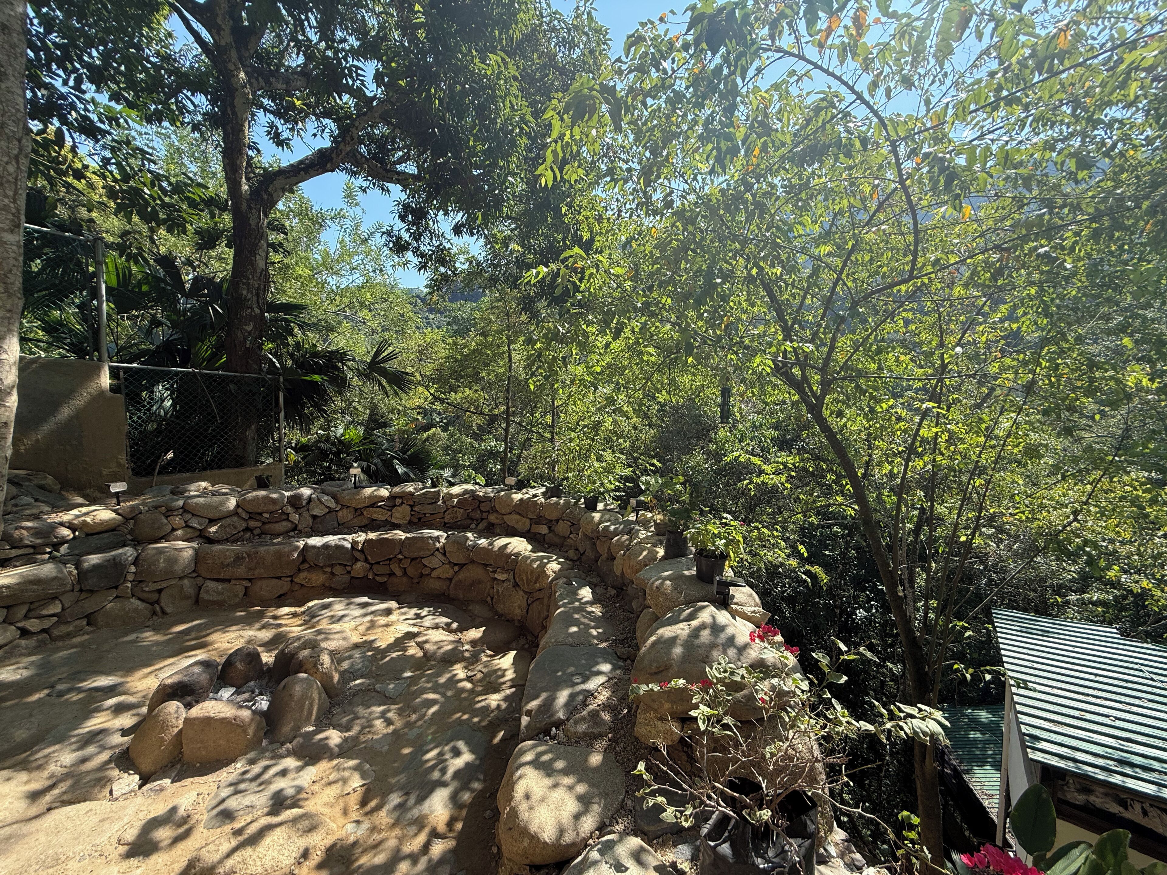 A stone wall enclosure with a fire pit, surrounded by trees and a metal roof structure.