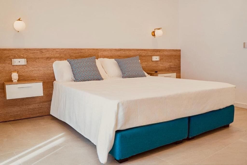 A hotel room with a wooden headboard, a large bed with a white bedspread, two bedside tables with lamps, and a blue upholstered bench.