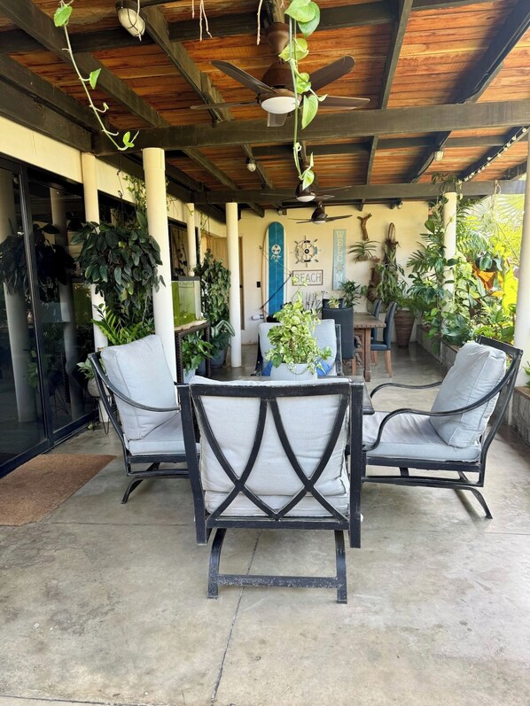 Un patio exterior con mesa y sillas, ventiladores de techo y diversas plantas en macetas.