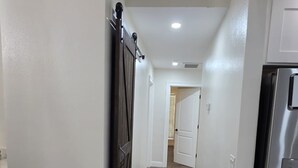 A hallway with a wooden floor, white walls, a door on the left, and a refrigerator on the right.