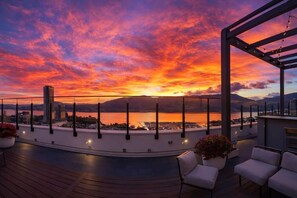Une terrasse sur le toit offrant une vue sur un coucher de soleil se reflétant sur une étendue d’eau, le tout entouré d’immeubles.