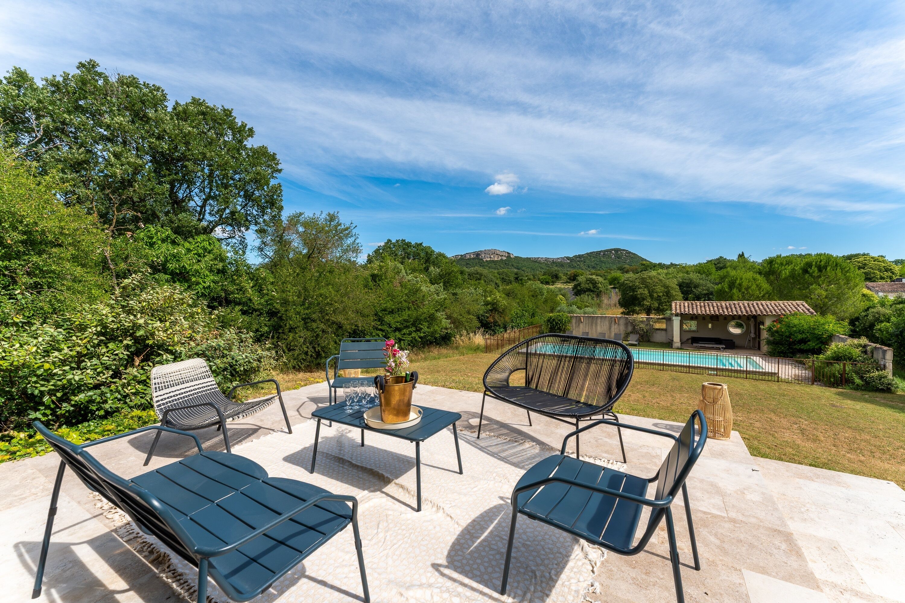 Une terrasse extérieure avec des chaises et une table en métal, un vase décoratif, et une vue sur une piscine et des collines en arrière-plan.