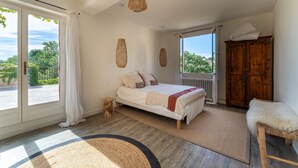 Ein Schlafzimmer mit einem Bett, einem Holzschrank, einem kleinen Hocker und Blick auf Grünflächen.