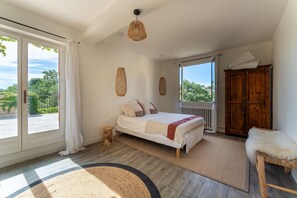 A bedroom with a bed, a wooden wardrobe, a small stool, and a view of greenery outside. - Grand'maison - Avec piscine privée (Bouquet)