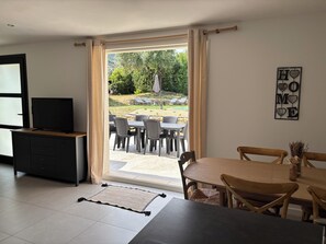 Ein Zimmer mit Fernseher auf einer Holzkommode, einem Esstisch mit Stühlen, einer Couch und Blick auf eine Terrasse mit Stühlen und Sonnenschirmen.