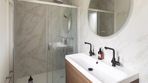 A bathroom with a glass-enclosed shower, a white sink with a wooden vanity, and a round mirror above the sink. - Casa Fiorenza - À 750 mètres de la plage (Dinard)