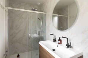A bathroom with a glass-enclosed shower, a white sink with a wooden vanity, and a round mirror above the sink. - Casa Fiorenza - À 750 mètres de la plage (Dinard)