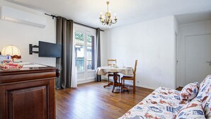A room with a wooden floor, a dining table with chairs, a sofa, a TV, and a chandelier. - L'Écrin Thermal - Appt avec balcon et vue dégagée (Amélie-les-Bains-Palalda)
