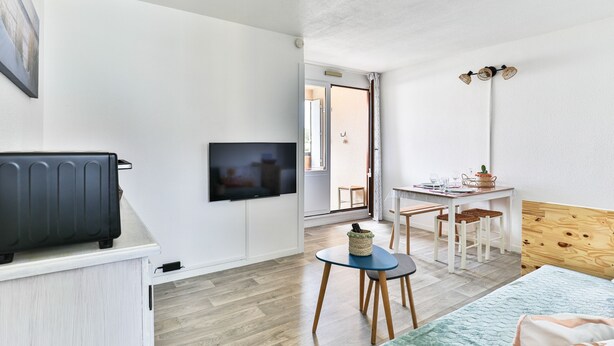 Ein Zimmer mit einem Bett, einem Fernseher, einem kleinen Tisch mit Stühlen, einem hölzernen Kopfteil und einer Tür zu einem weiteren Raum.