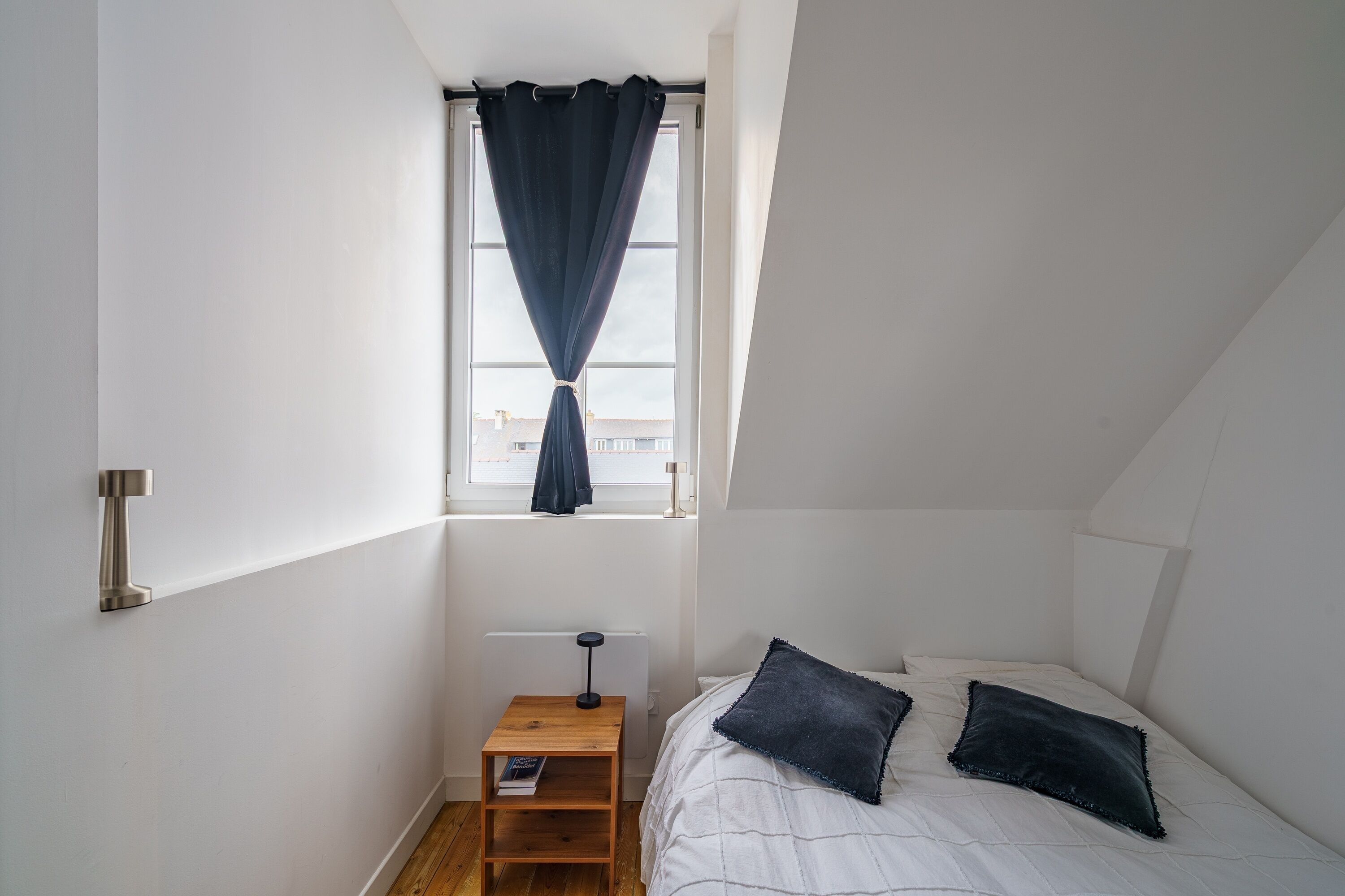 A bedroom with a bed, two pillows, a nightstand with a lamp, a window with curtains, and a radiator.
