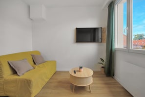 Une pièce avec un canapé jaune, une petite table ronde en bois sur laquelle est posée une plante, un téléviseur fixé au mur et une fenêtre avec des rideaux.