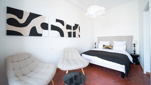 Quarto com cama, duas cadeiras, uma mesinha e arte abstrata nas paredes.