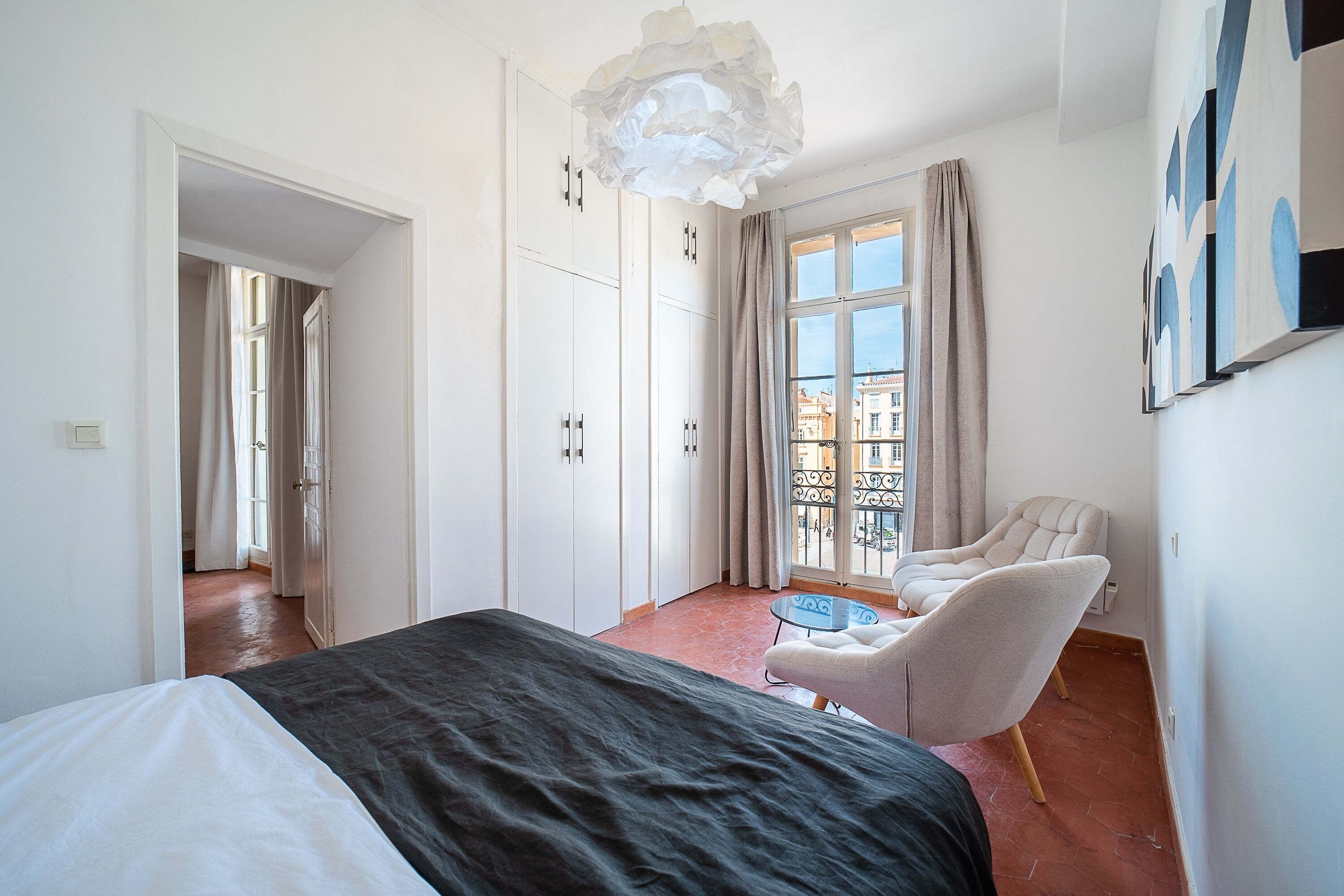 A bedroom with a bed, a chair, a table, a balcony with a view, and a large window.
