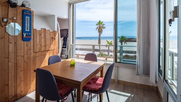 A room with a wooden dining table, chairs, a large window with a view of the beach, and a wooden wall with a mirror and a blue sign. - Escale à Canet - Au bord de la plage (Canet-en-Roussillon)