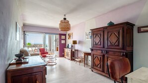 A spacious room with a large wooden cabinet, a dining table with chairs, a wicker chair, and a view through a glass door. - Escale à Canet - Au bord de la plage (Canet-en-Roussillon)