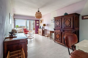 A spacious room with a large wooden cabinet, a dining table with chairs, a wicker chair, and a view through a glass door. - Escale à Canet - Au bord de la plage (Canet-en-Roussillon)