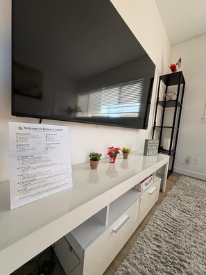 Ein modernes Wohnzimmer mit einem an der Wand befestigten Flachbildfernseher, einer weißen Unterhaltungsecke mit einem Schild und einem kleinen Regal mit Dekoration.