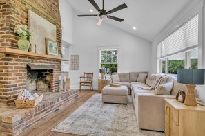 A living room with a brick fireplace, a ceiling fan, a large window, and a sectional sofa. - Evergreen Cottage Escape (Pegram)