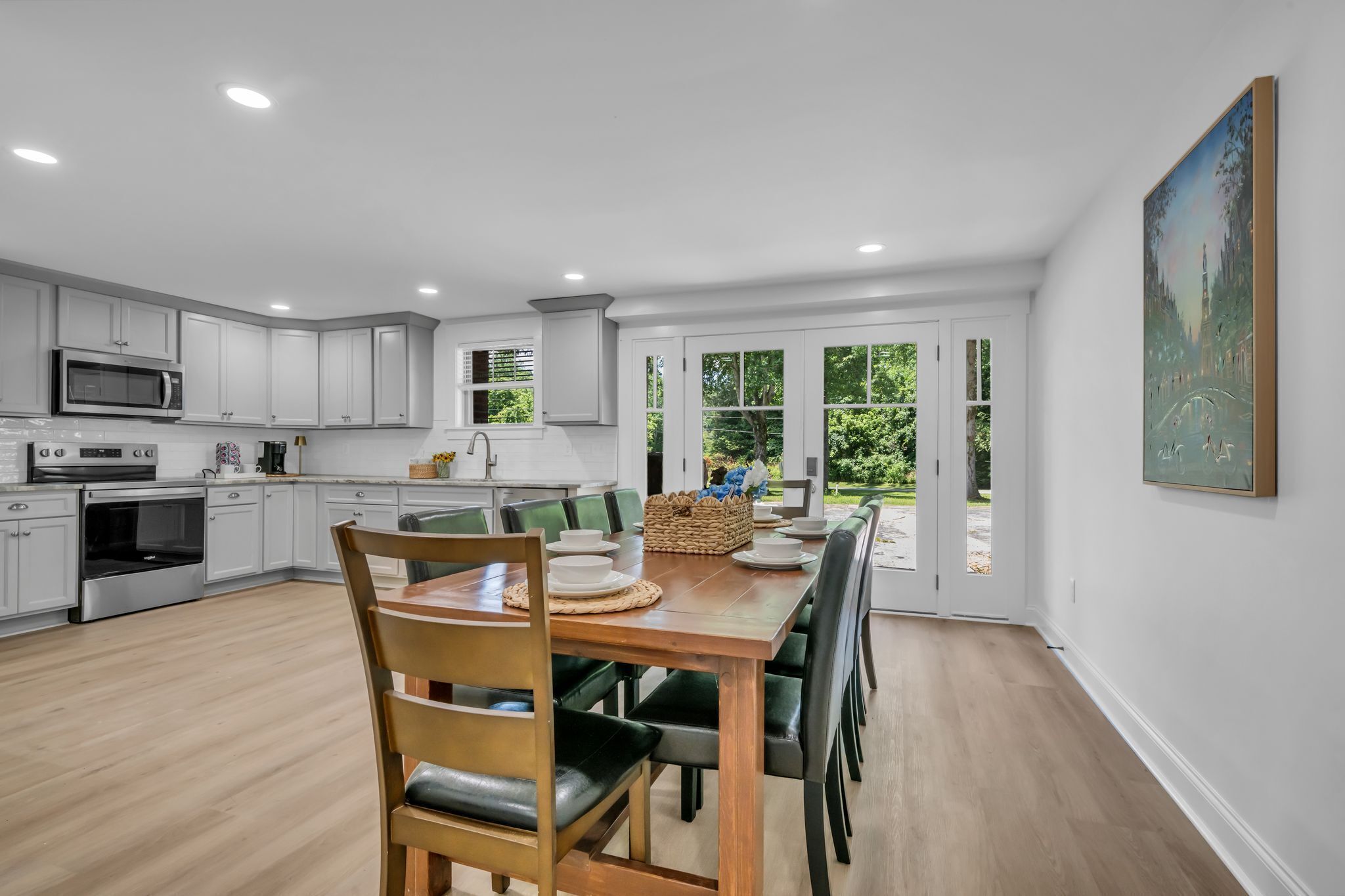 A modern kitchen with white cabinets, a dining table set for six, and a large window overlooking greenery.