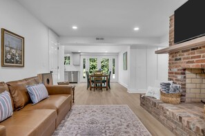 A modern living room with a brown leather sofa, a patterned rug, a dining area with a table and chairs, and a brick fireplace. - Evergreen Cottage Escape (Pegram)