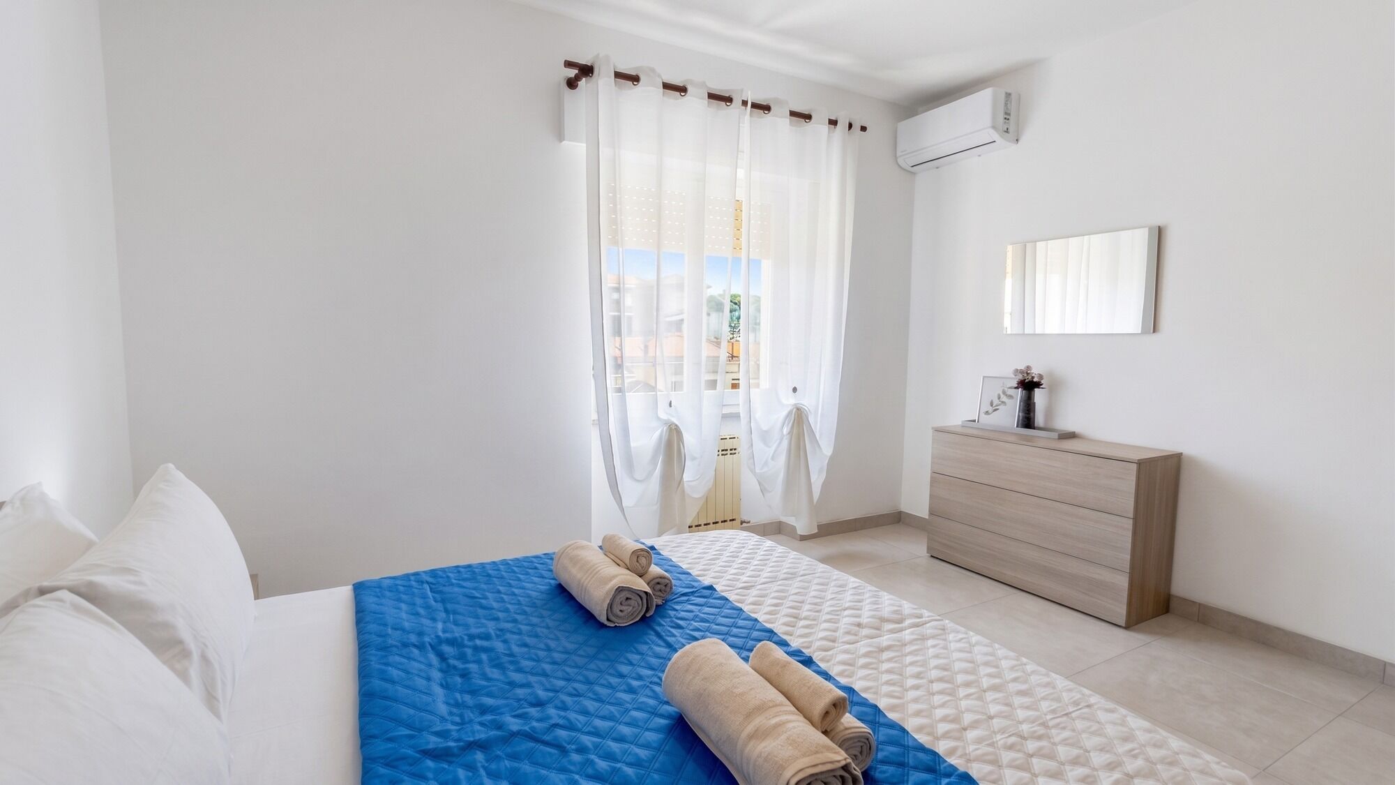 A hotel room with a bed, a dresser, a mirror, and a window with sheer curtains.
