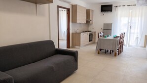 Een moderne woonkamer met een bank, een eettafel gedekt voor vier personen, een keuken met houten kastjes, en een flatscreen televisie aan de muur.