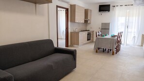 A modern living room with a sofa, a dining table set for four, a kitchen with wooden cabinets, and a flat-screen TV mounted on the wall. - Urban Nest Alghero - Two Bedroom Apartment, Sleeps 5 (Alghero)