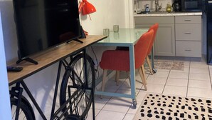 Espai habitable compacte amb televisió sobre un prestatge de fusta, taula de dinar amb caires vermelles, zona de cuina amb armaris i electrodomèstics, i una catifa amb dibuixos al sòl.