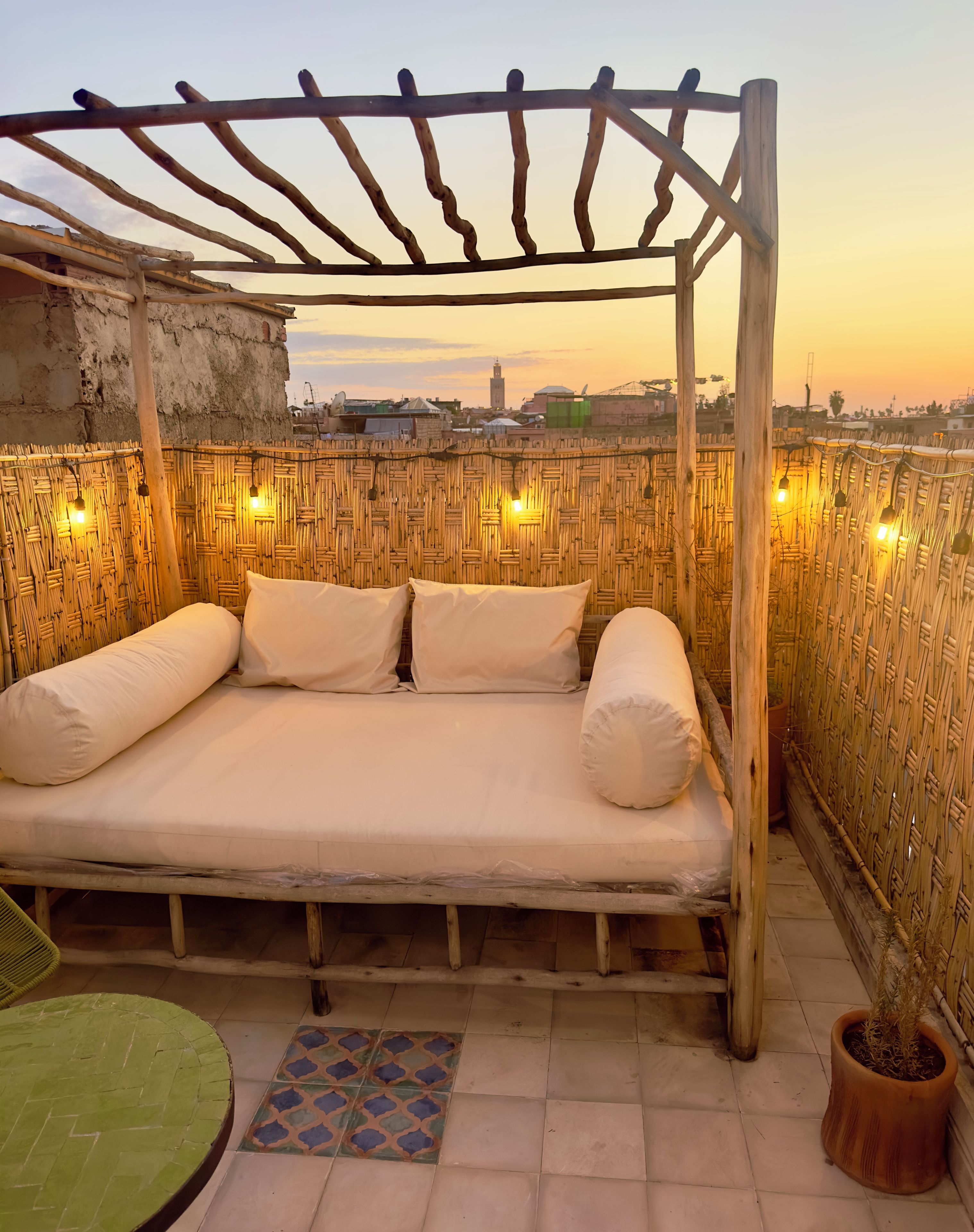 A bed with a wooden frame and white bedding, set against a backdrop of a woven wall and a cityscape at sunset.