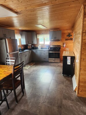 A compact kitchen with wooden cabinets, a stainless steel refrigerator, a stove, and a dining area with a table and chairs. - Lovely 3-bedroom cabin with AC in cool Longville (Longville)