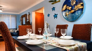 Un comedor con una mesa dispuesta para cuatro personas, rodeada de sillas de mimbre, una pared azul decorada con peces y estrellas de mar, y una puerta que da a otra habitación.