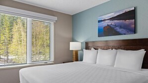 Une chambre d’hôtel avec un grand lit, une fenêtre avec des stores, une lampe de chevet et un tableau accroché au mur.