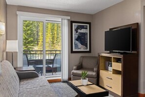 Ein Wohnzimmer mit Sofa, Couchtisch, Fernseher und Bücherregal. Ein Fenster bietet Blick auf Bäume.